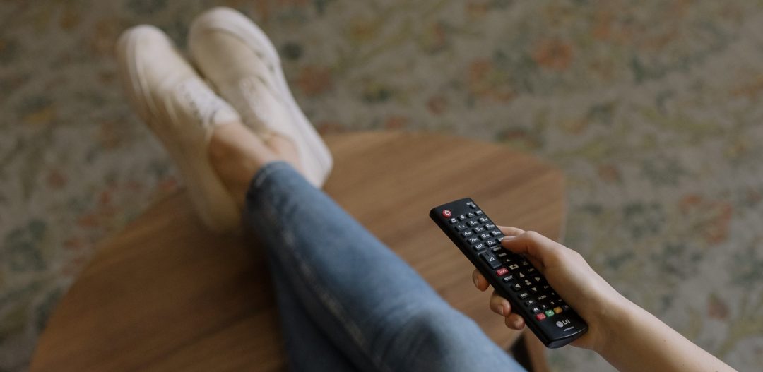 Woman's legs crossed wearing jeans and white sneakers on a wood table. Holding out a remote in hand. Floral carpet