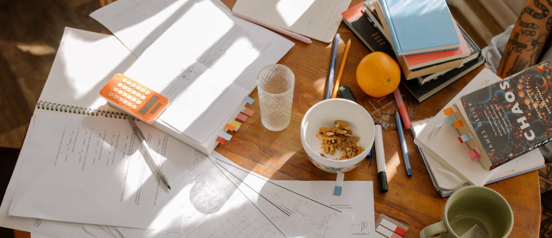 wood table with scattered papers, pens, books, and coffee mugs